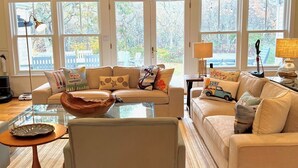 A cozy living room with a sofa, a coffee table, and a glass-top table. There are large windows with a view of the outside. - Secluded, Private Vacation Beach Home on Chappaquiddick Island, Walk To Beach. (Edgartown)