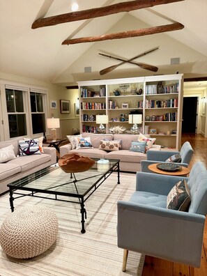 A spacious living room with a high ceiling, exposed wooden beams, a large bookshelf, a comfortable sofa set, and a coffee table.