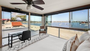 Habitació moderna amb una finestra gran que dona a una platja i un port esportiu, ventilador de sostre i escriptori amb cadira.