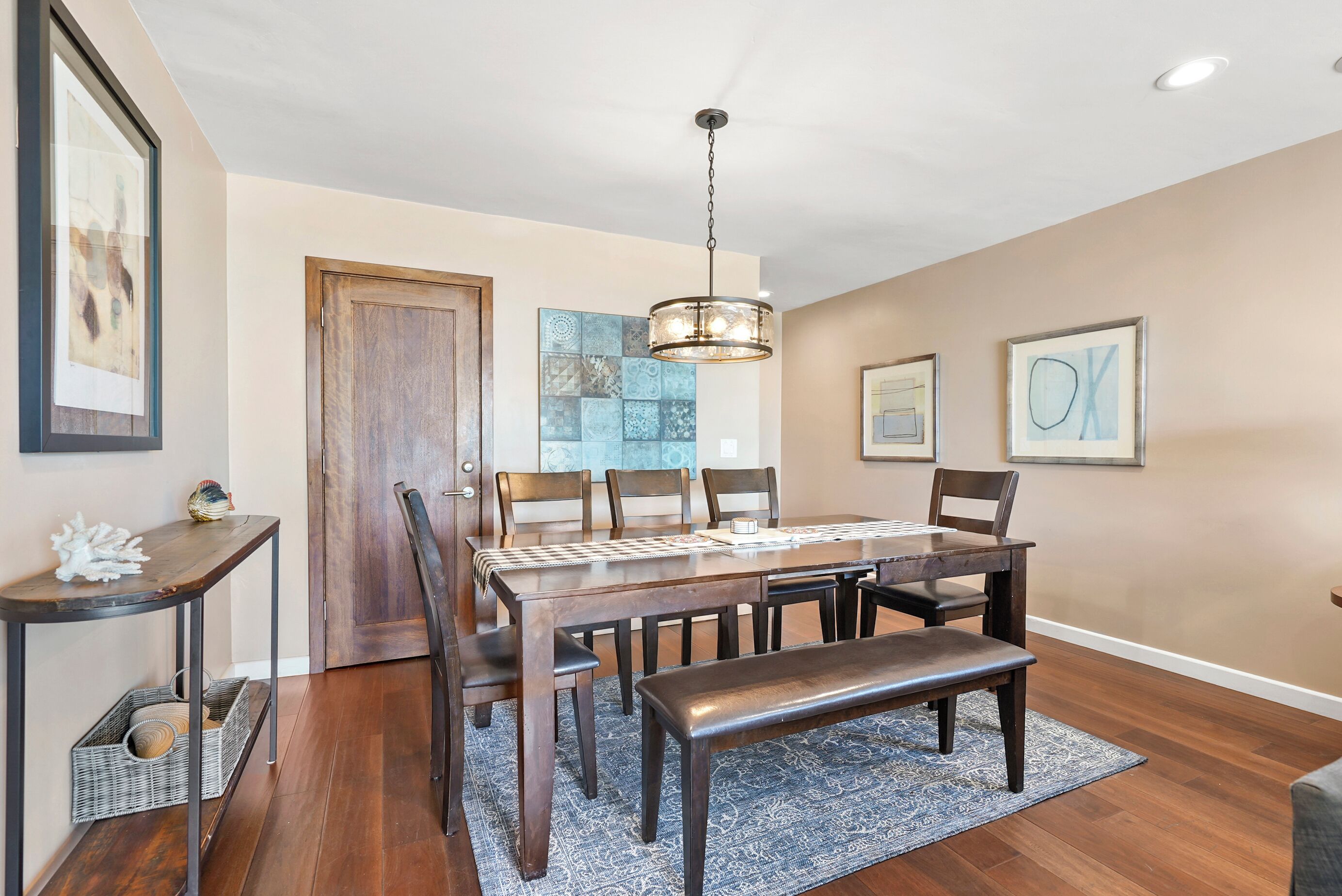 A dining area with a wooden table, chairs, a bench, a hanging light fixture, and a patterned rug.