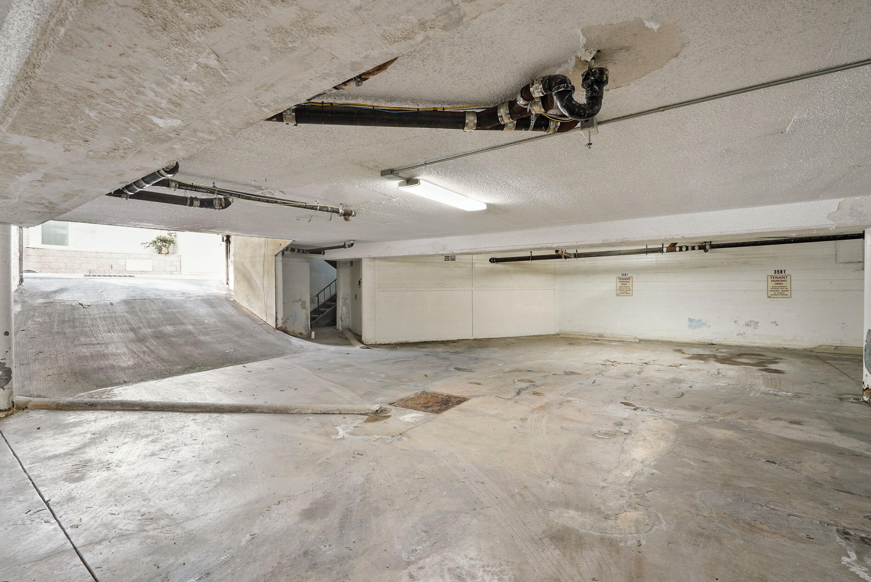 Een lege kamer met een betonnen vloer, zichtbare leidingen in het plafond en een deur naar een andere ruimte.