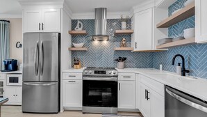 A modern kitchen with white cabinetry, a stainless steel refrigerator, a built-in oven, and a herringbone-patterned backsplash. - Panoramic oceanfront views, ground floor luxury renovation next to Ocean Annie’s (Myrtle Beach)