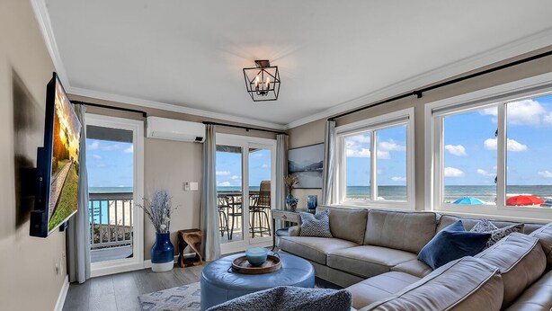 A spacious living room with a large sofa, a coffee table, and a TV mounted on the wall. There are multiple windows providing views of the beach and ocean. - Panoramic oceanfront views, ground floor luxury renovation next to Ocean Annie’s (Myrtle Beach)