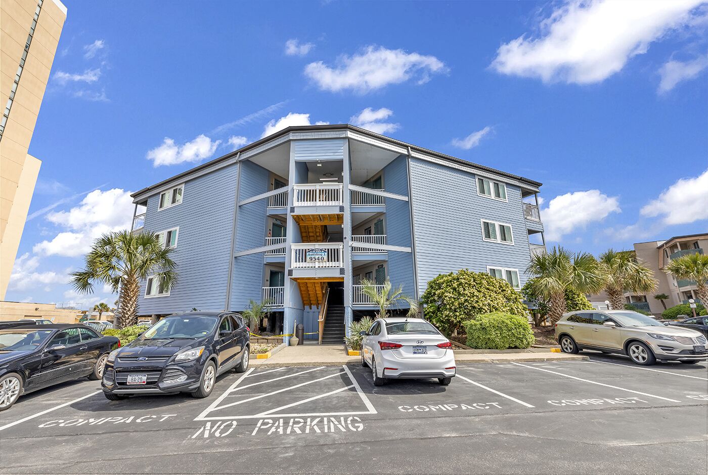 A multi-story building with a blue exterior, a balcony on the second floor, and a parking lot with 'NO PARKING' and 'COMPACT' signs.