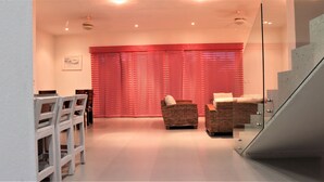 En moderne stue med en glas trappe, et spiseområde med hvide stole og et stort vindue med røde gardiner.