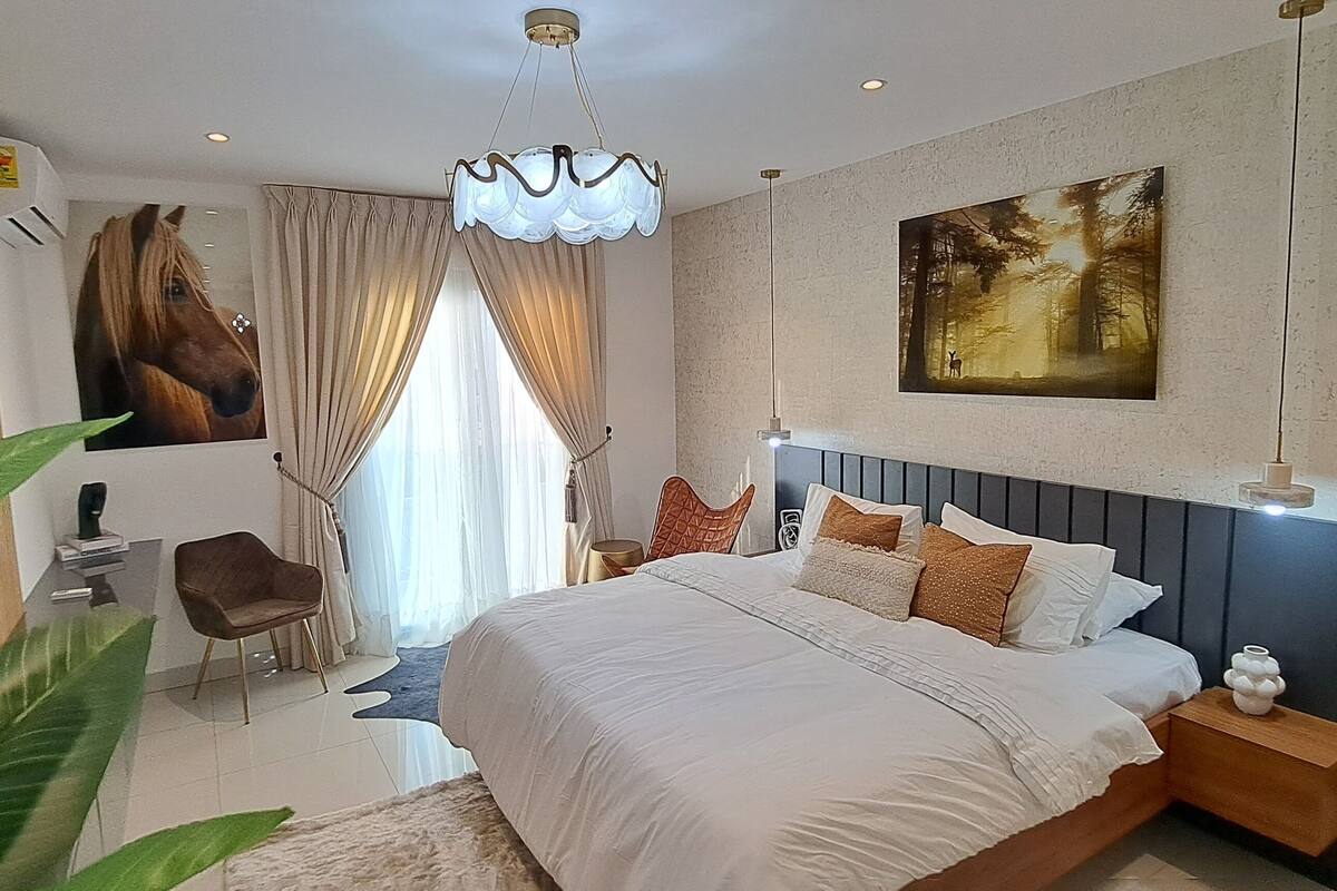 A modern bedroom with a large bed, a desk, and a chair. There is a painting on the wall and a plant in the corner.