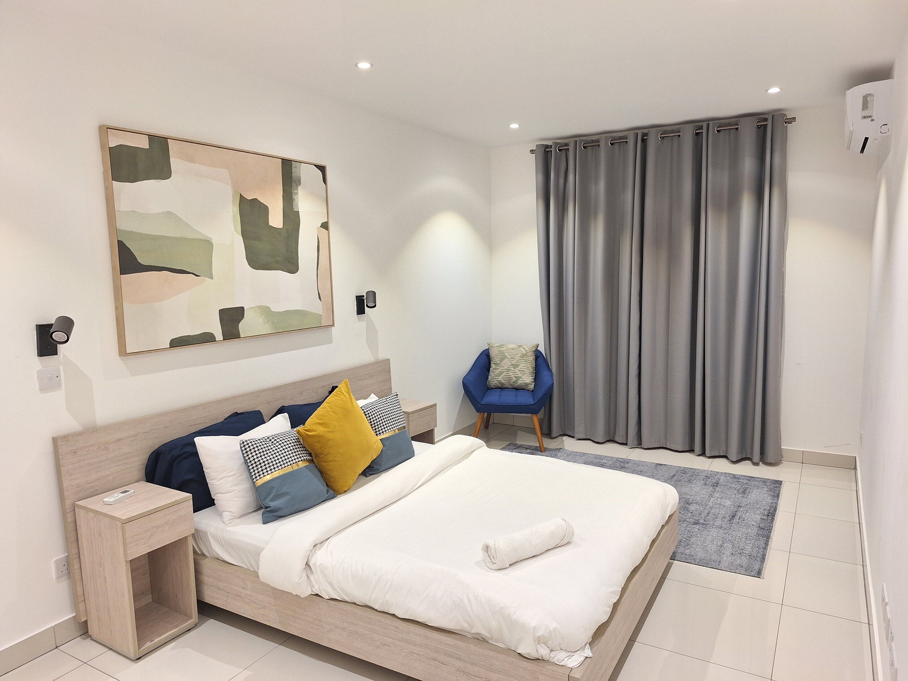 A modern hotel room with a neatly made bed, a side table, a blue armchair, a grey curtain, and a wall-mounted abstract painting.
