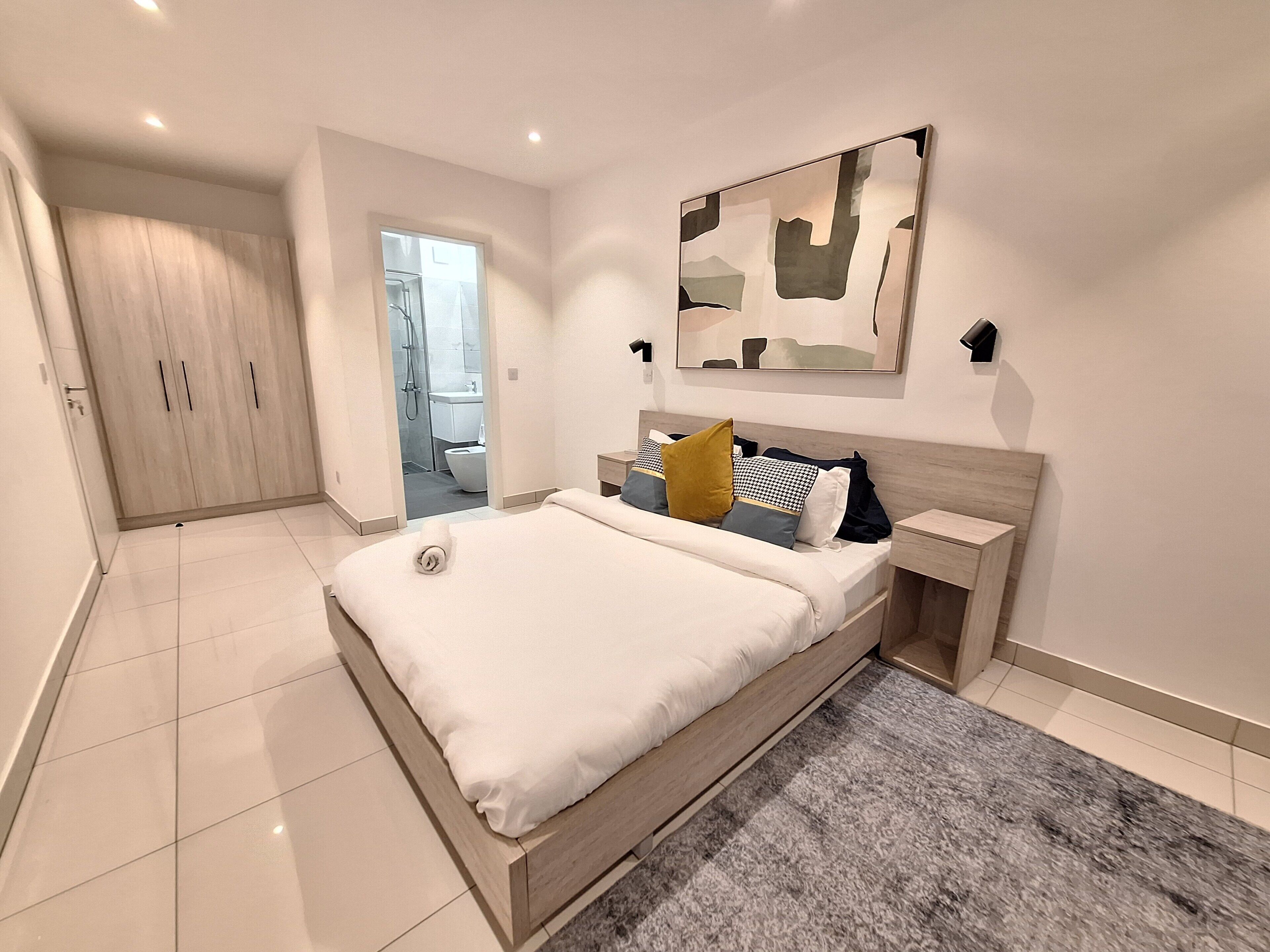 A modern bedroom with a large bed, a nightstand, a wall-mounted lamp, and a door leading to a bathroom.