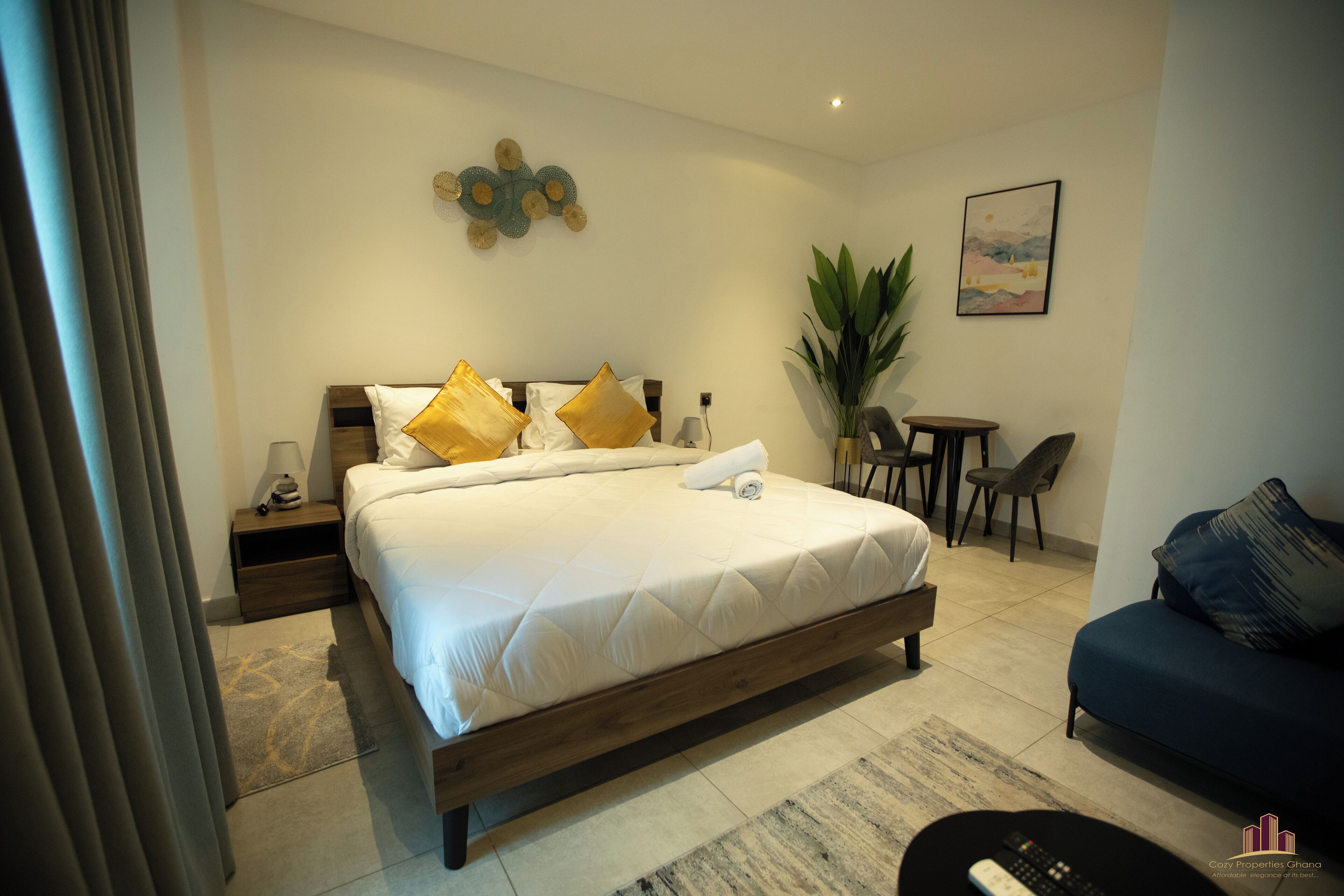 A hotel room with a bed, bedside tables, a desk, a chair, a sofa, a plant, and a painting on the wall.