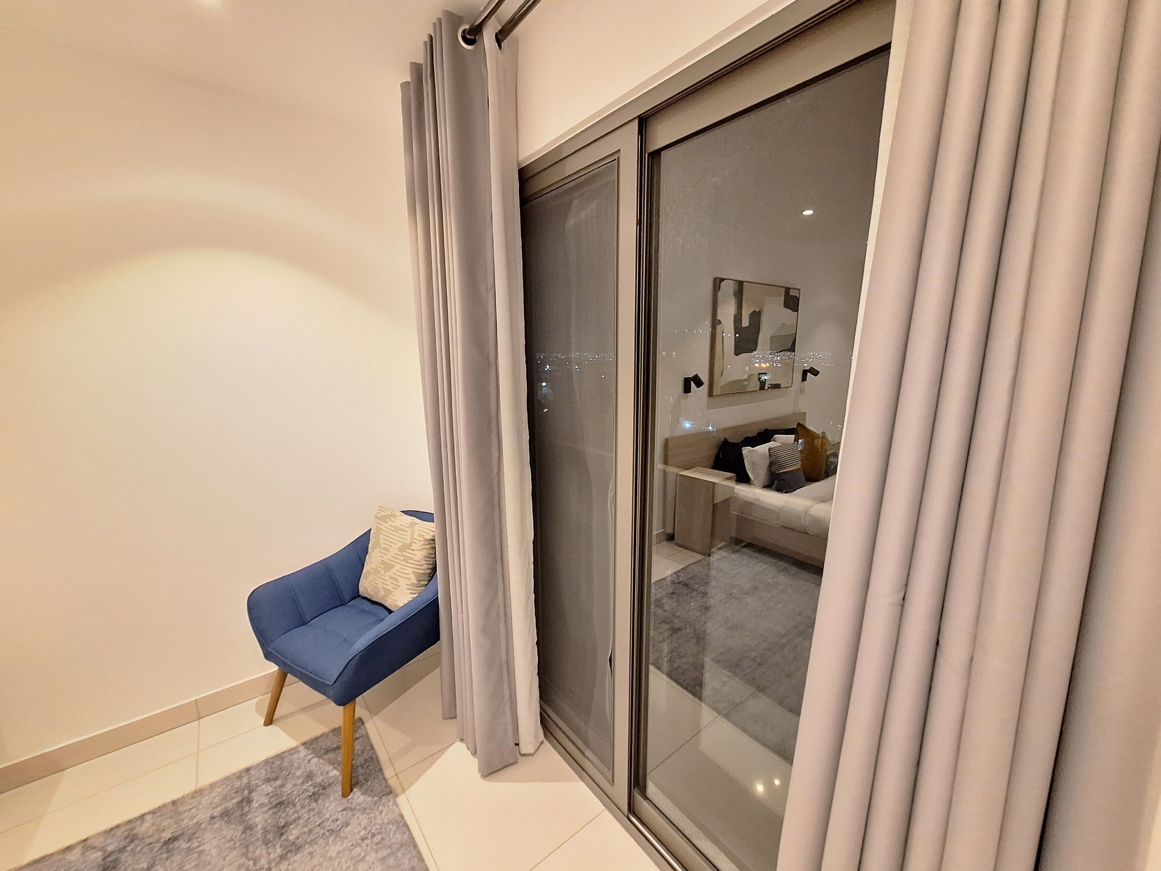 Une chambre moderne dotée d’une porte-fenêtre coulissante donnant sur un balcon, d’une chaise bleue et d’une table de chevet avec une lampe.