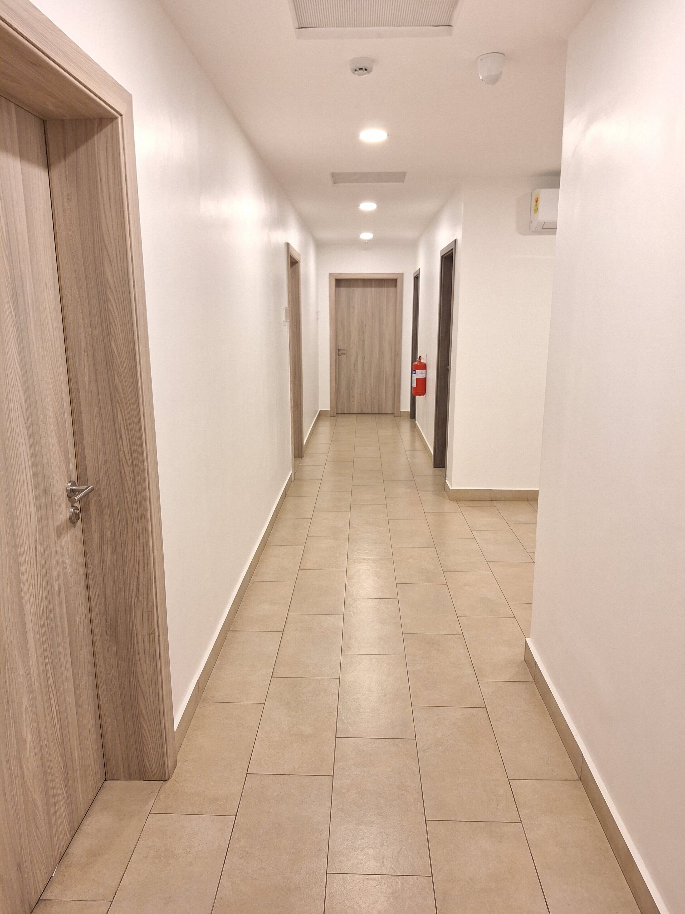 Ein sauberer, gut beleuchteter Korridor mit Holztüren und Fliesenboden, der zu einem Raum mit einer Holztür und einem an der Wand befestigten Feuerlöscher führt.