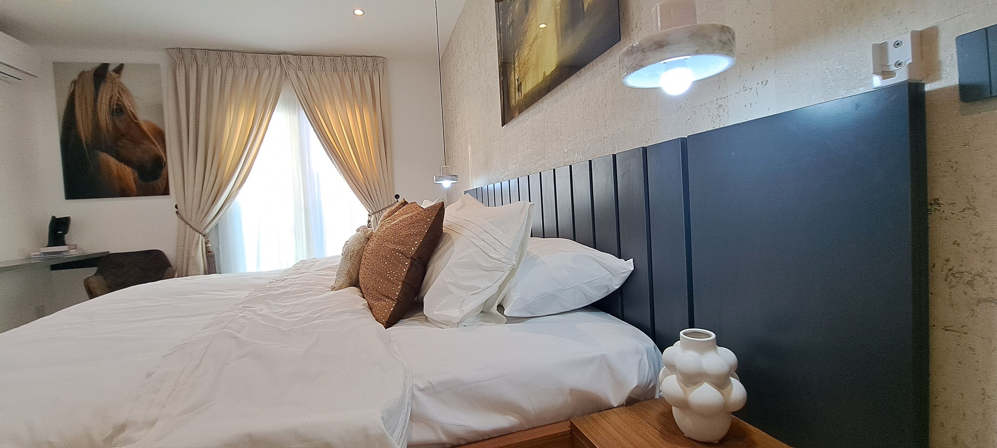 A bedroom with a large bed, beige curtains, a mounted painting, a bedside lamp, and a wall-mounted light fixture.