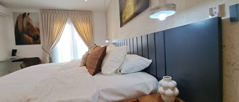 A bedroom with a large bed, beige curtains, a mounted painting, a bedside lamp, and a wall-mounted light fixture.