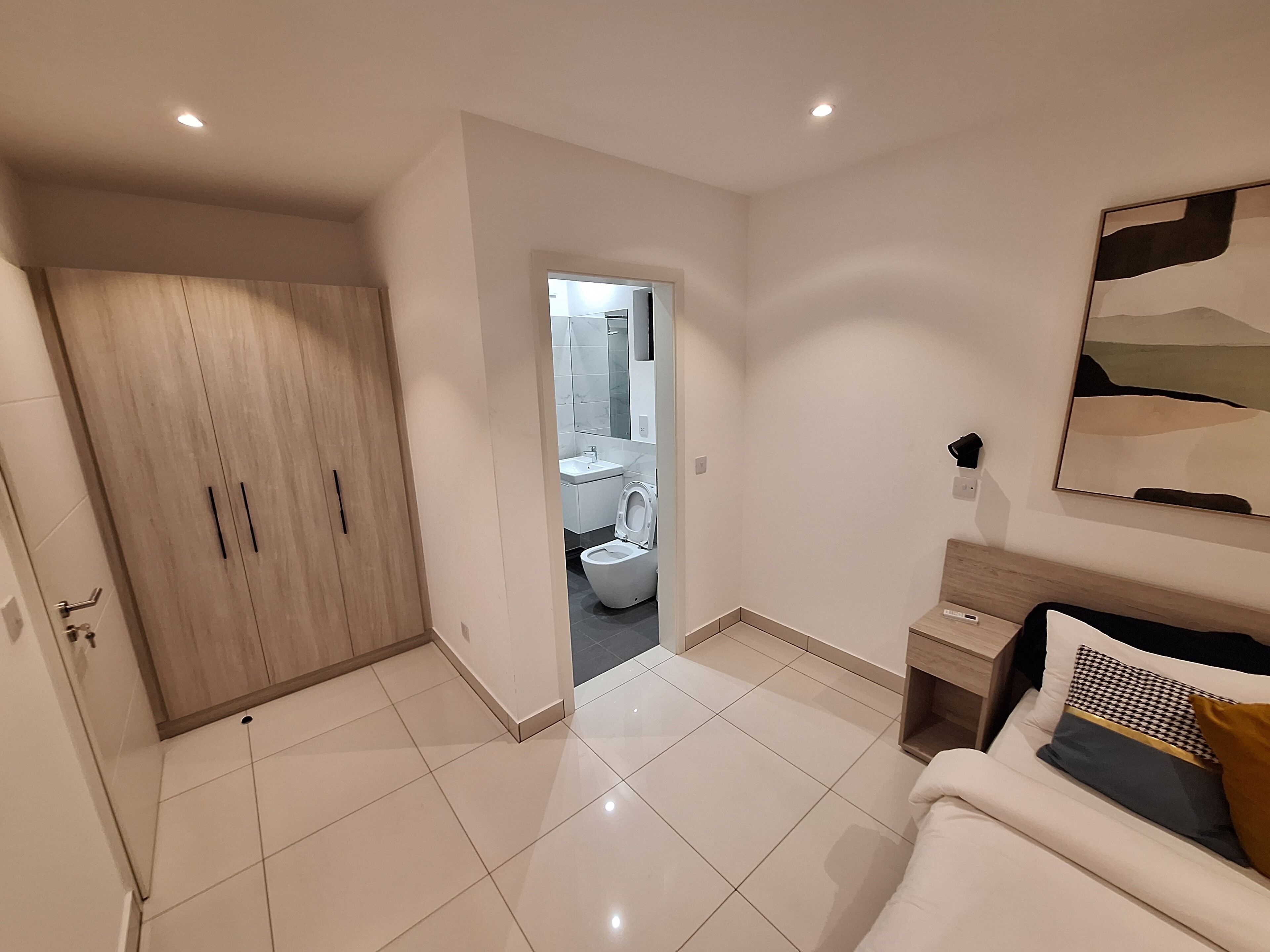 A modern bedroom with a large wardrobe, a bedside table, a bed with pillows, and a bathroom visible through an open door.