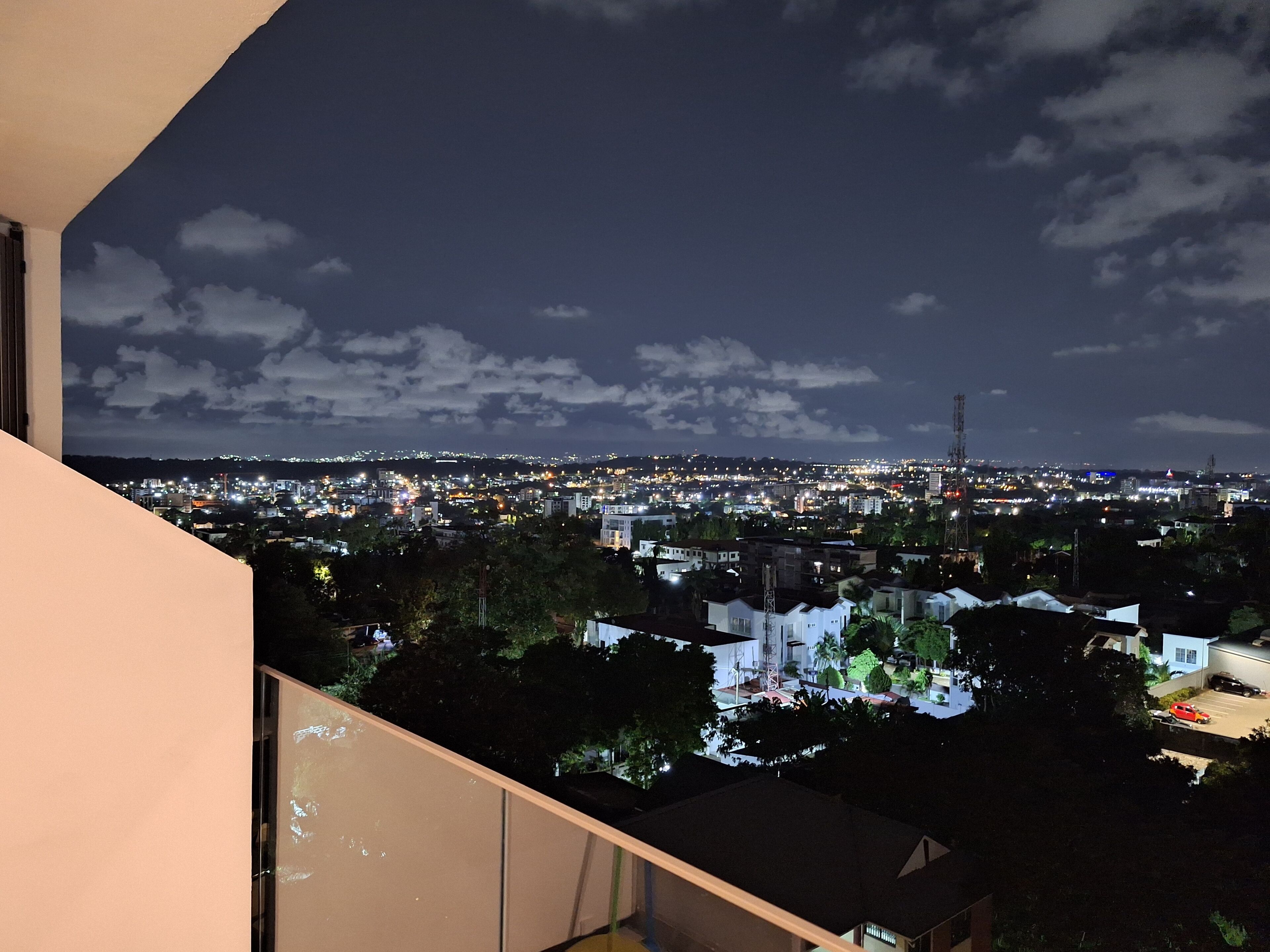 A nighttime cityscape viewed from a balcony with a clear sky and scattered clouds.