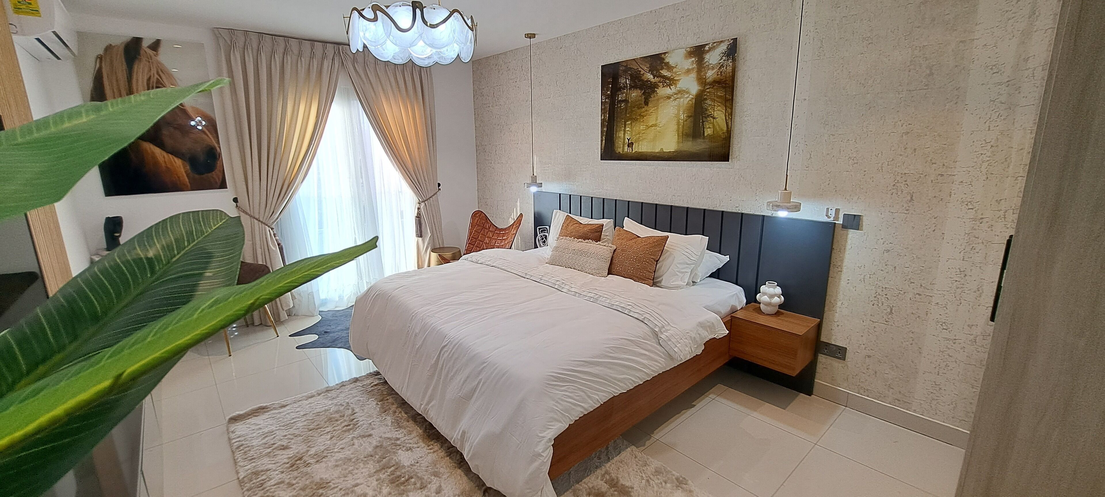 A modern bedroom with a large bed, a nightstand, a wall-mounted artwork, and a large window with curtains.