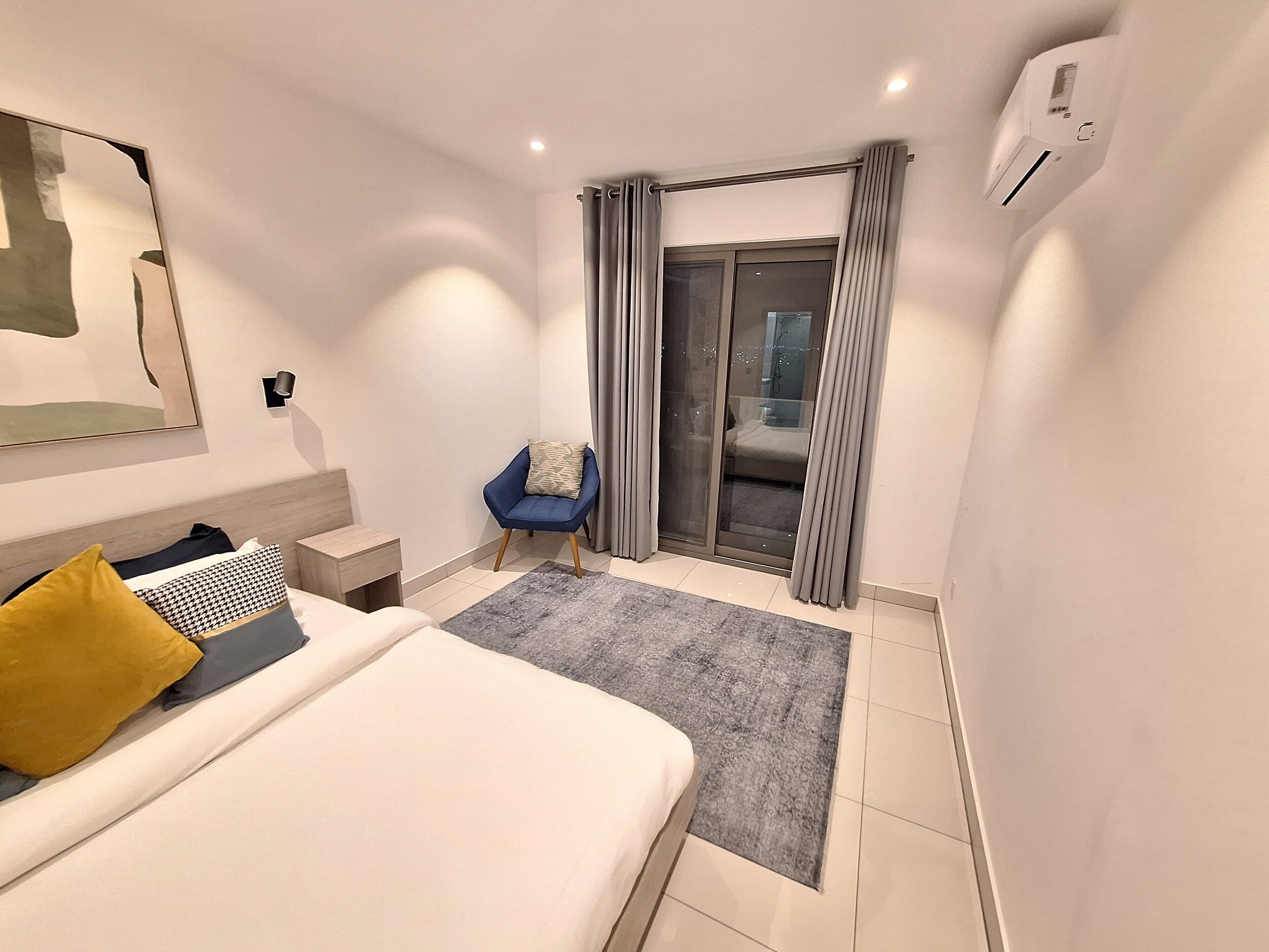 Een moderne slaapkamer met een groot bed, een klein nachtkastje, een blauwe stoel en een glazen schuifdeur naar een balkon.