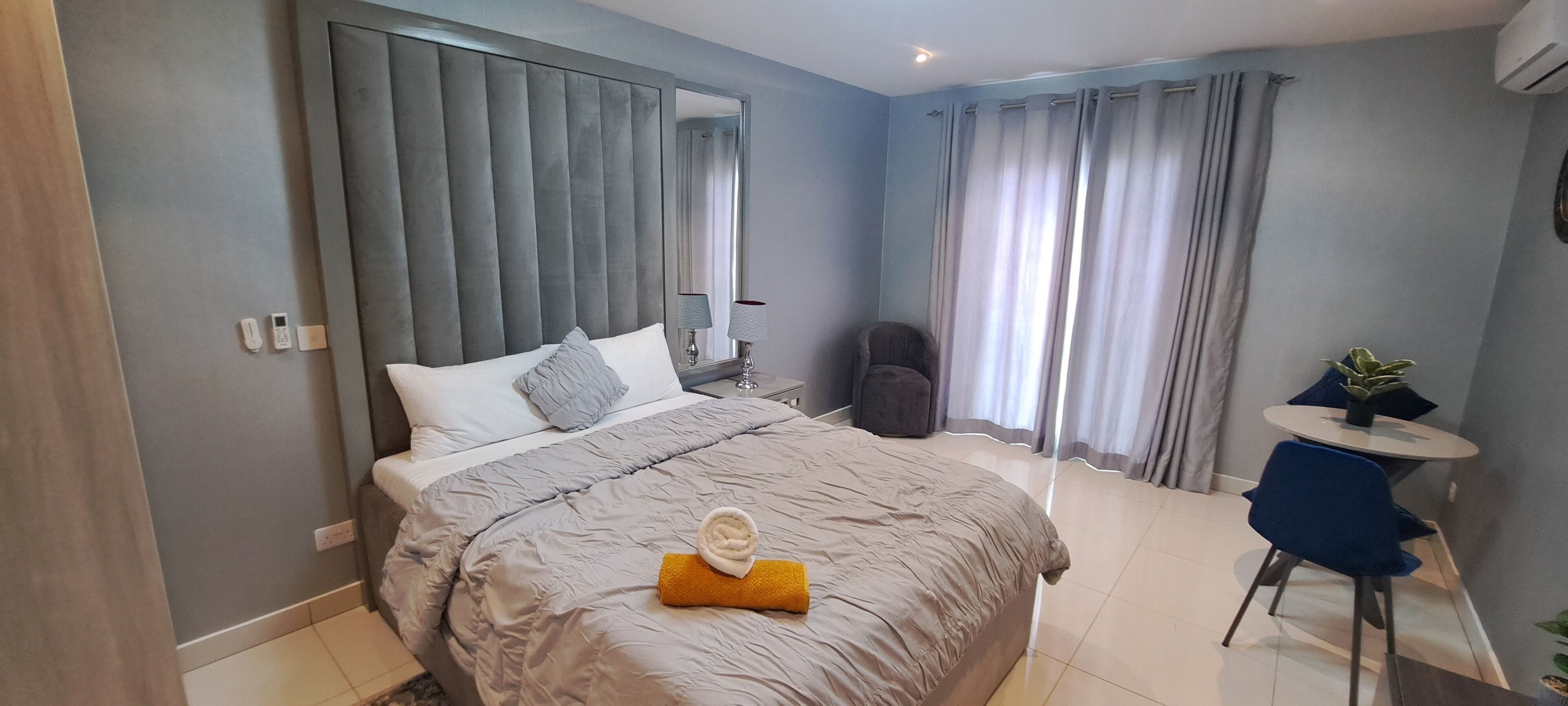 A modern hotel room with a large bed, a bedside table, a chair, a small round table, and a potted plant.