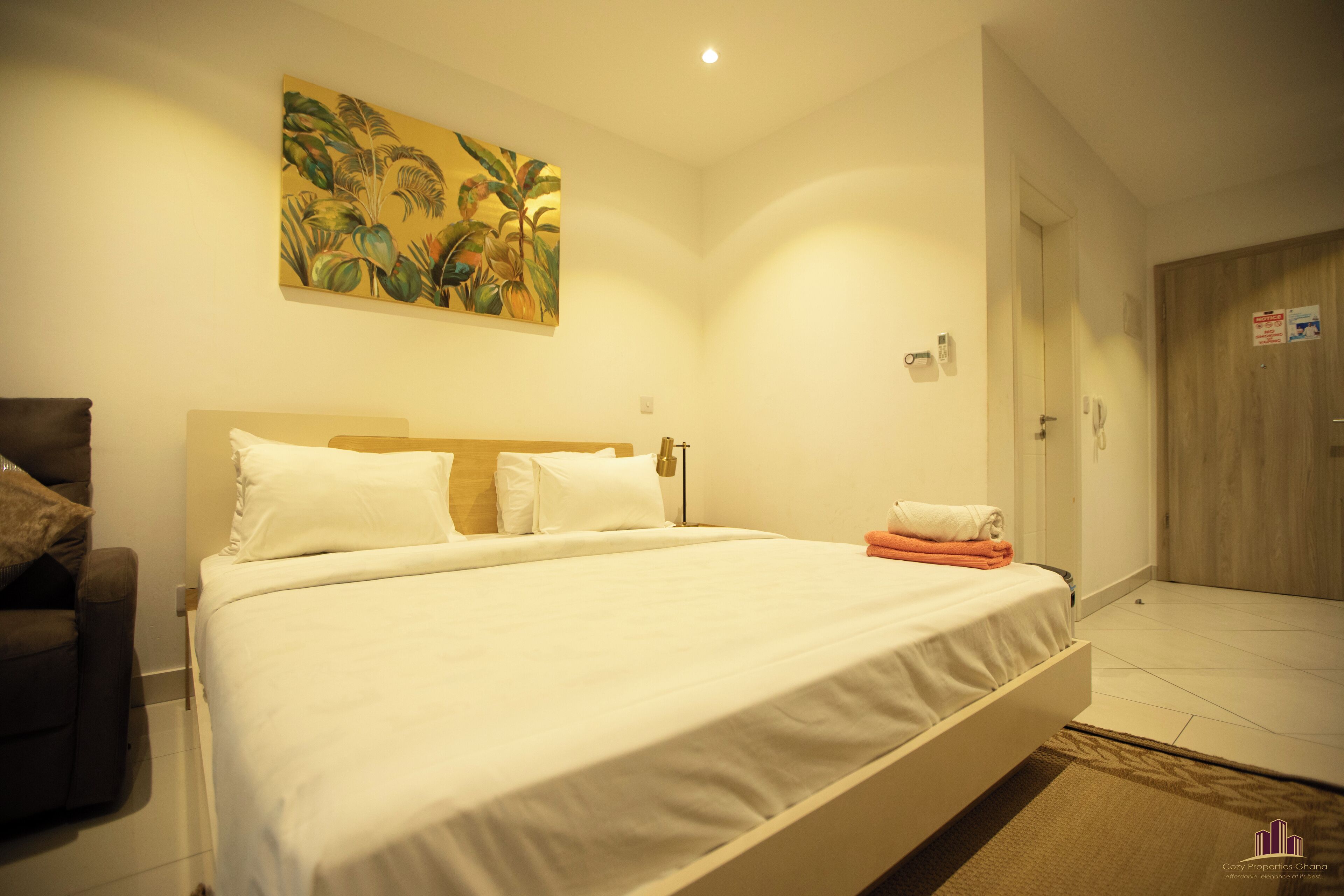 Habitació d'hotel amb un llit gran, un quadre a la paret, una porta de fusta i una taula petita amb una tovallola.