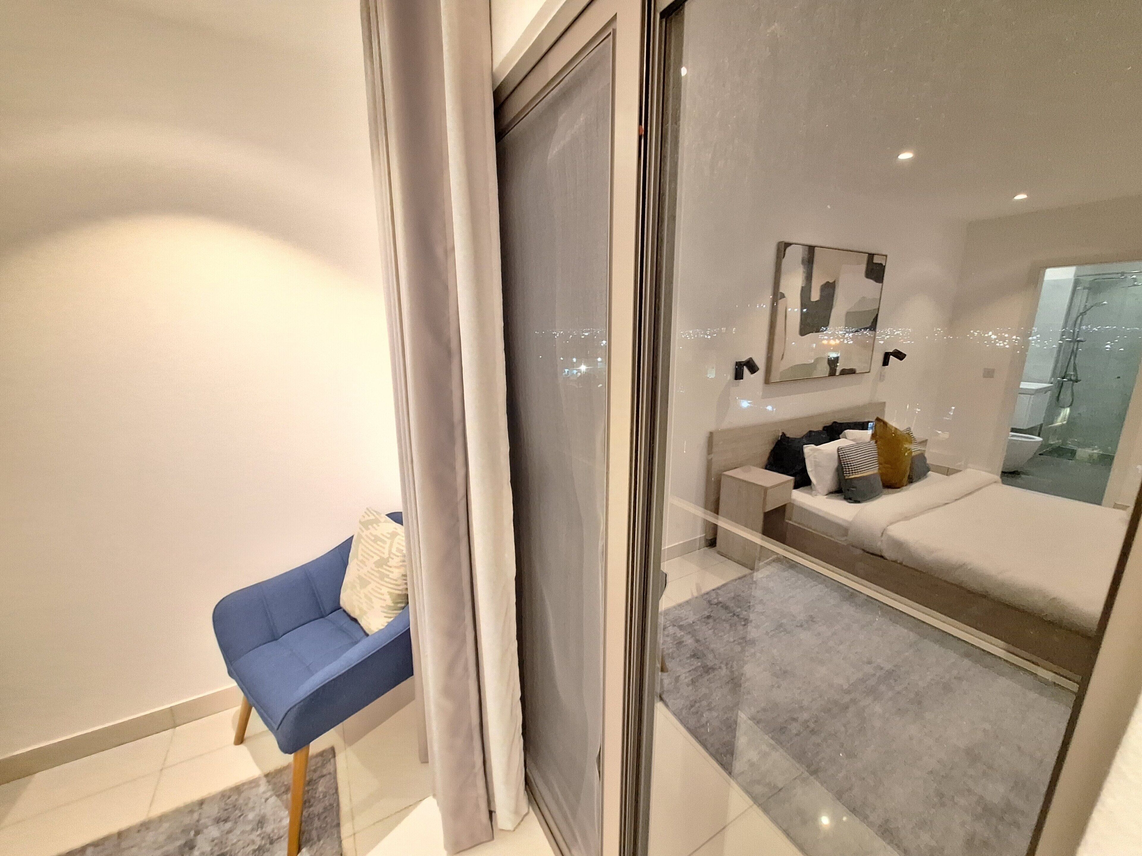 Ein modernes Schlafzimmer mit Glastür zum Badezimmer. Es gibt einen blauen Stuhl, ein Bett mit Kopfteil und einen Spiegel an der Wand.