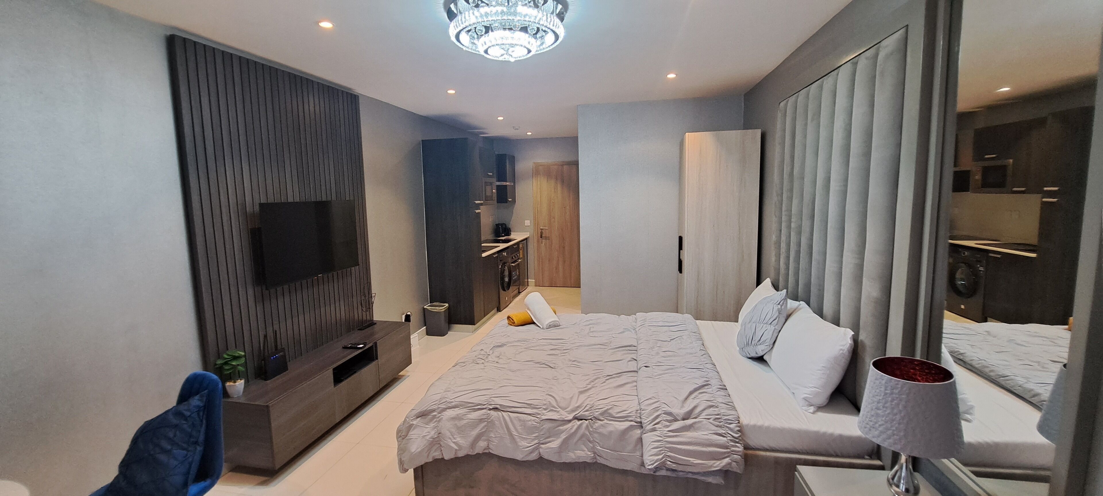 A modern bedroom with a large bed, a TV mounted on the wall, a bedside table, and a mirror.