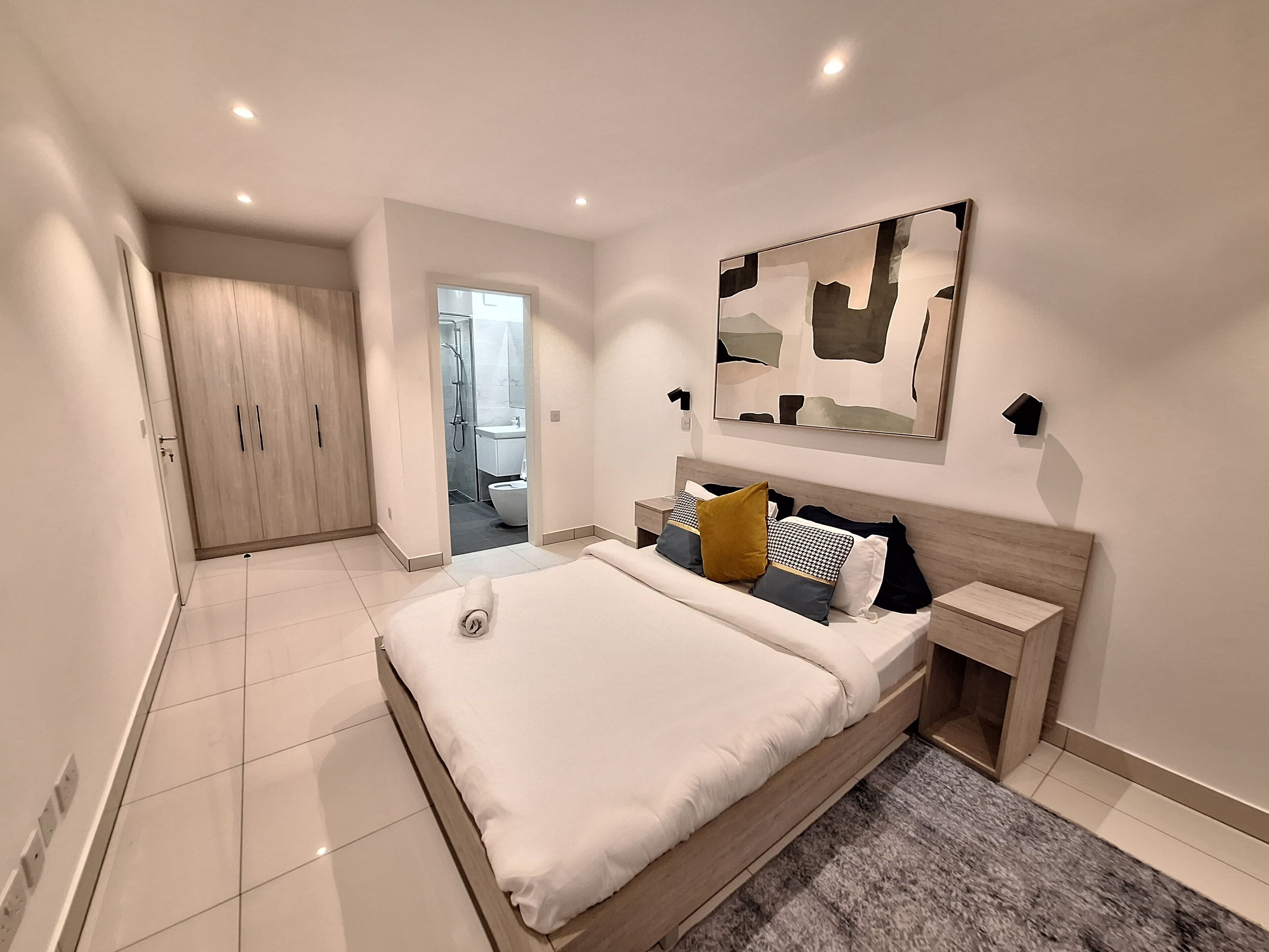 A modern bedroom with a large bed, a wooden wardrobe, a bathroom visible through an open door, and a contemporary abstract painting on the wall.