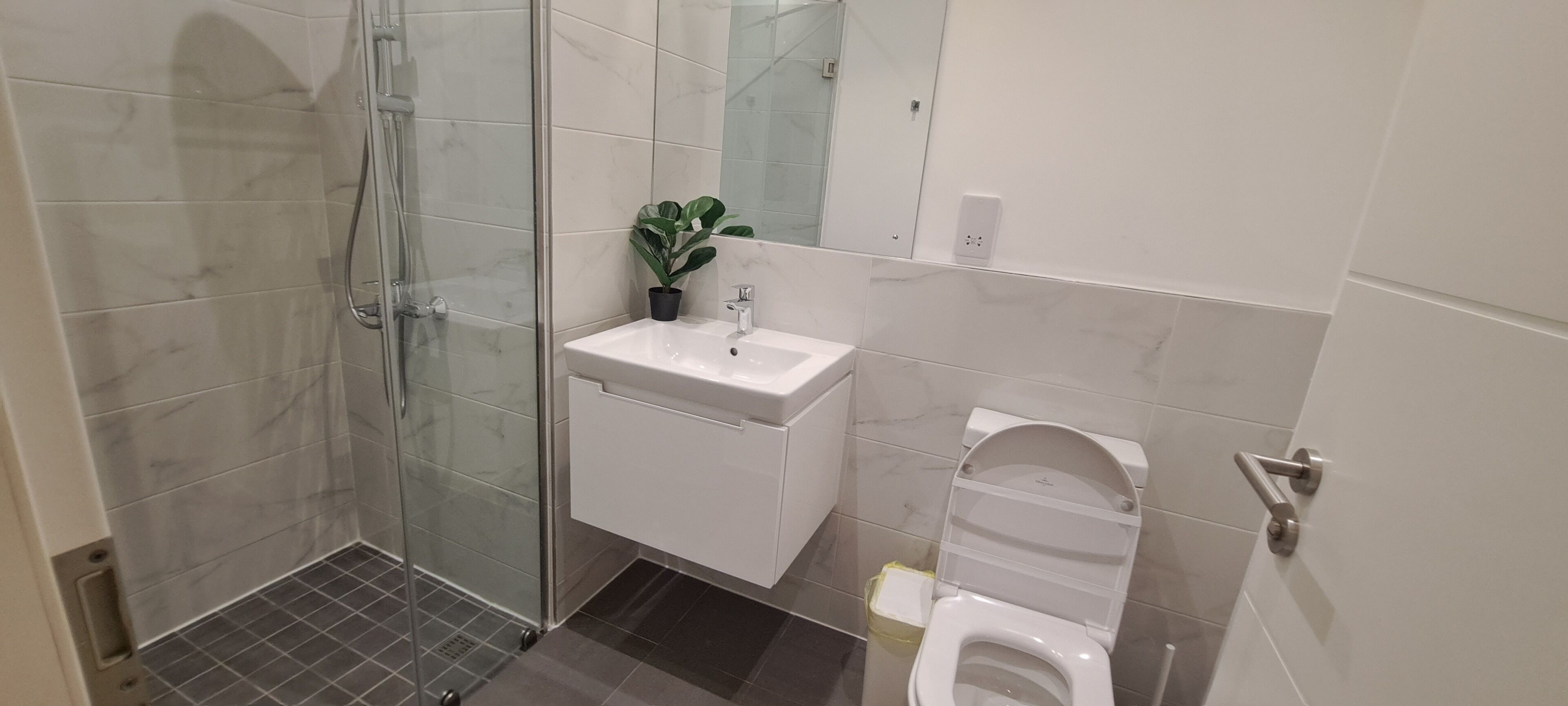 Een badkamer met een douchecabine van glas, een witte wastafel, een plant in pot en een toilet.