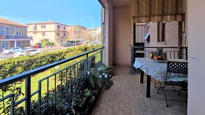 En balkon med et bord og stole, plantekrukker og udsigt til en gade med bygninger og parkerede biler.