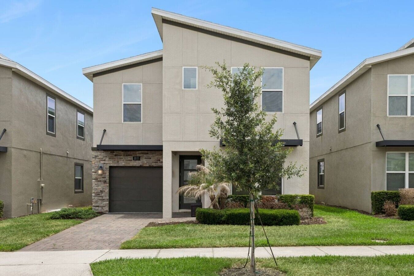 A modern two-story residential building with a garage, a small tree in the front yard, and a well-maintained lawn.