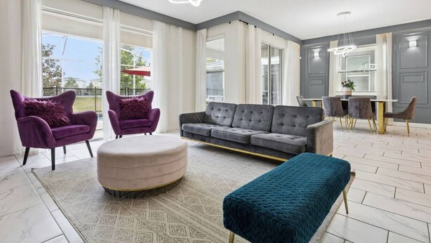 A spacious living room with a grey sofa, two purple armchairs, a round ottoman, and a teal bench.