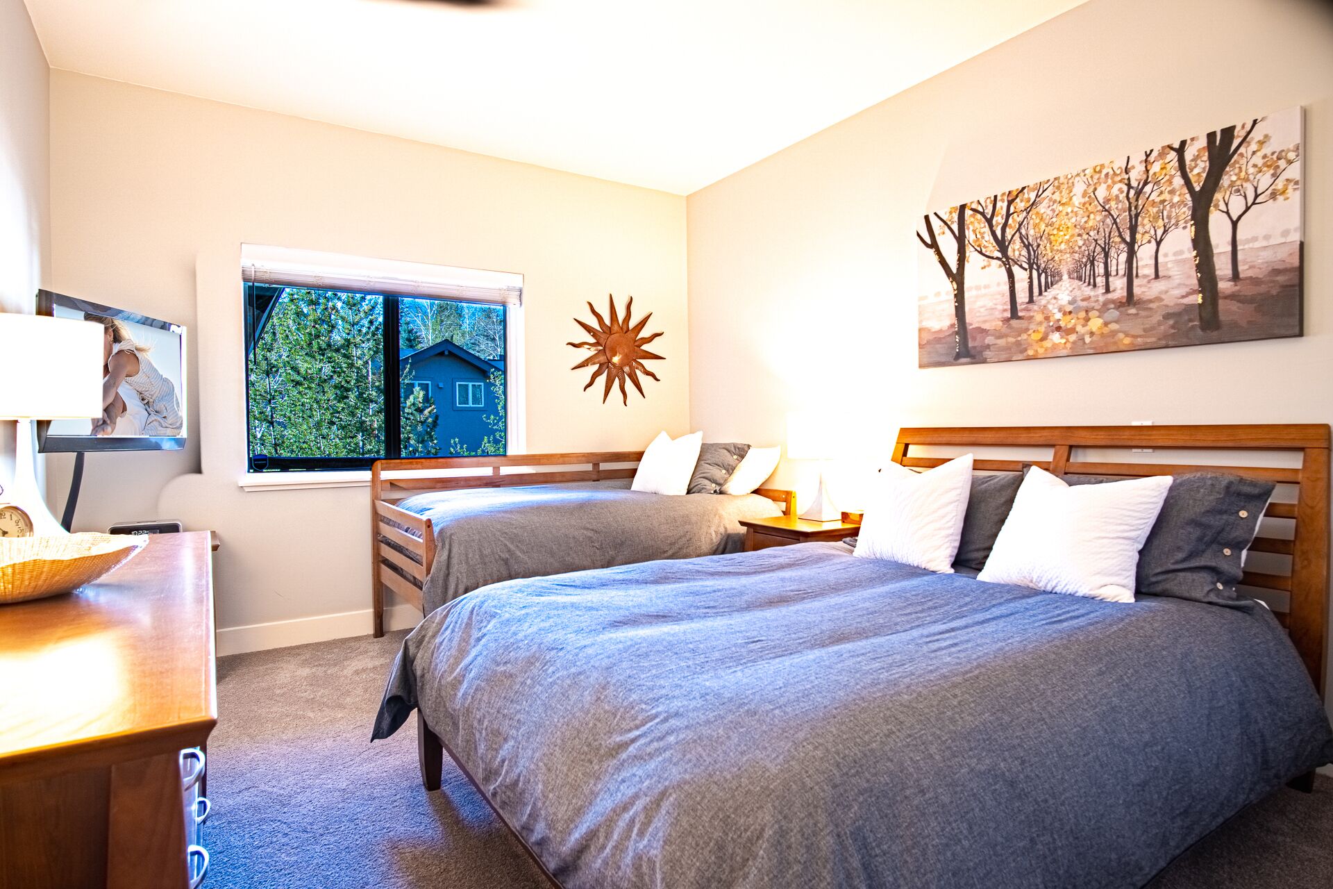 A bedroom with two beds, a window with a view of greenery, a sun decoration, and a painting of trees.