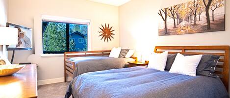 A bedroom with two beds, a window with a view of greenery, a sun decoration, and a painting of trees.