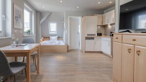 Moderni hotellihuone, jossa on puulattia, valkoisilla lakanoilla varustettu sänky, ruokailutila pöydän ja tuolien kera sekä pieni keittiö kaappeineen ja televisiineen.