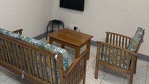 Een kamer met een plafondventilator, een flatscreen televisie aan de muur, een houten tafel, twee houten stoelen en twee ingelijste schilderijen.
