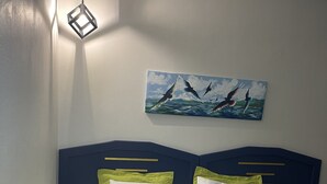 Een net opgemaakt bed met een groene deken en kussens, een schilderij van vogels aan de muur en een hangende lamp.