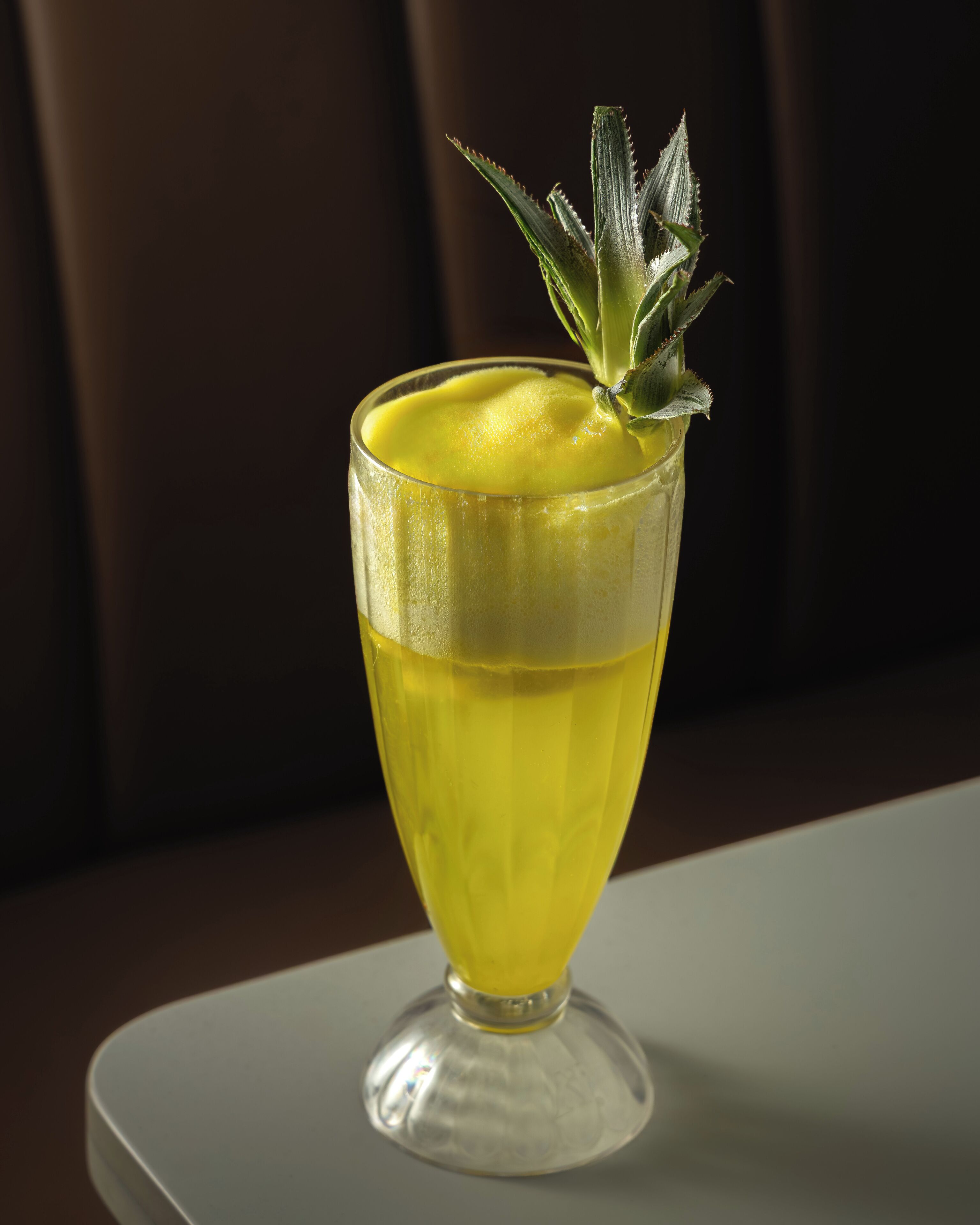 Un vaso con una bebida amarilla, una cuña de lima y una ramita de menta sobre una mesa blanca.