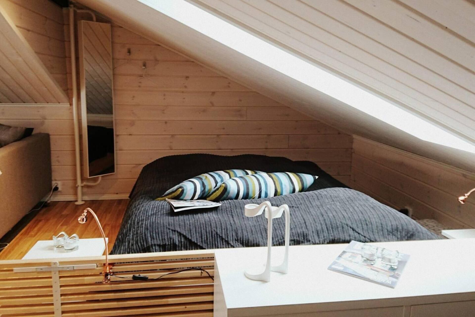 Et lille, hyggeligt soveværelse på loftet med skråvægge, en seng med stribede puder, et sengebord med en lampe og et spejl på væggen.