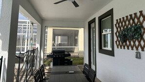 Un patio couvert avec un ventilateur de plafond, un barbecue et une vue sur une maison, visible à travers une porte vitrée.