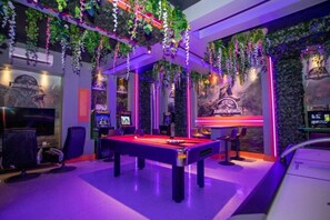 Ein Billardtisch mit Queue, Stühle und einem Neon-Schild mit der Aufschrift 'Jurassic World' in einem Raum mit hängenden Pflanzen und Spielautomaten.