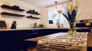 Una cocina moderna con armarios oscuros, encimeras de madera y una isla central. Hay estantes sobre el mostrador con vasos y un reloj. Un jarrón con flores está sobre la isla.