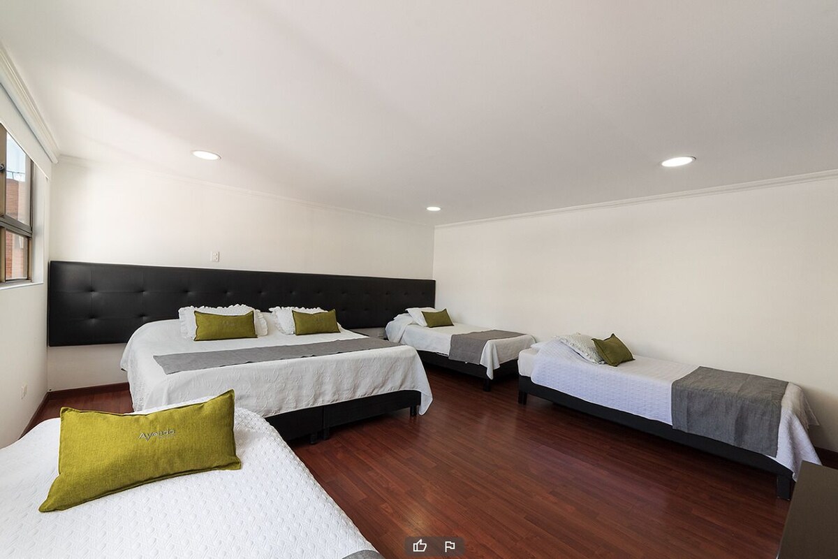 A hotel room with three beds arranged in a row, a large black headboard, and a window on the left side.