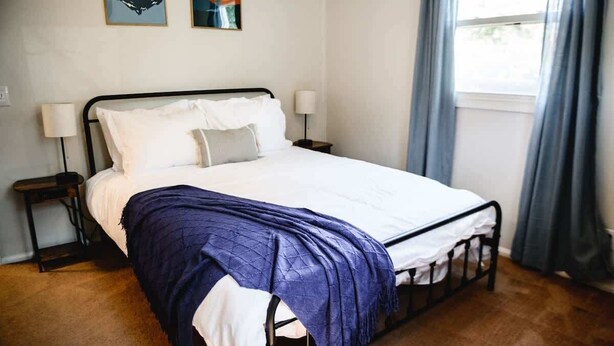 A neatly made bed with a white duvet and a blue blanket, two bedside lamps, and a window with curtains. - Urban Cottage:Homey & Convenient (Huntington)