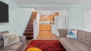 A living room with a sofa, a coffee table, and a rug. There is a staircase leading to a kitchen with wooden cabinets and a refrigerator. - The Battery Harbour Lower Suite (St. John's)
