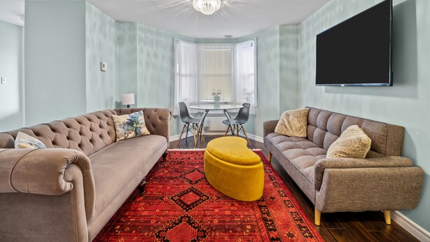 A living room with two sofas, a yellow ottoman, a chandelier, and a flat-screen TV on the wall. - The Battery Harbour Lower Suite (St. John's)