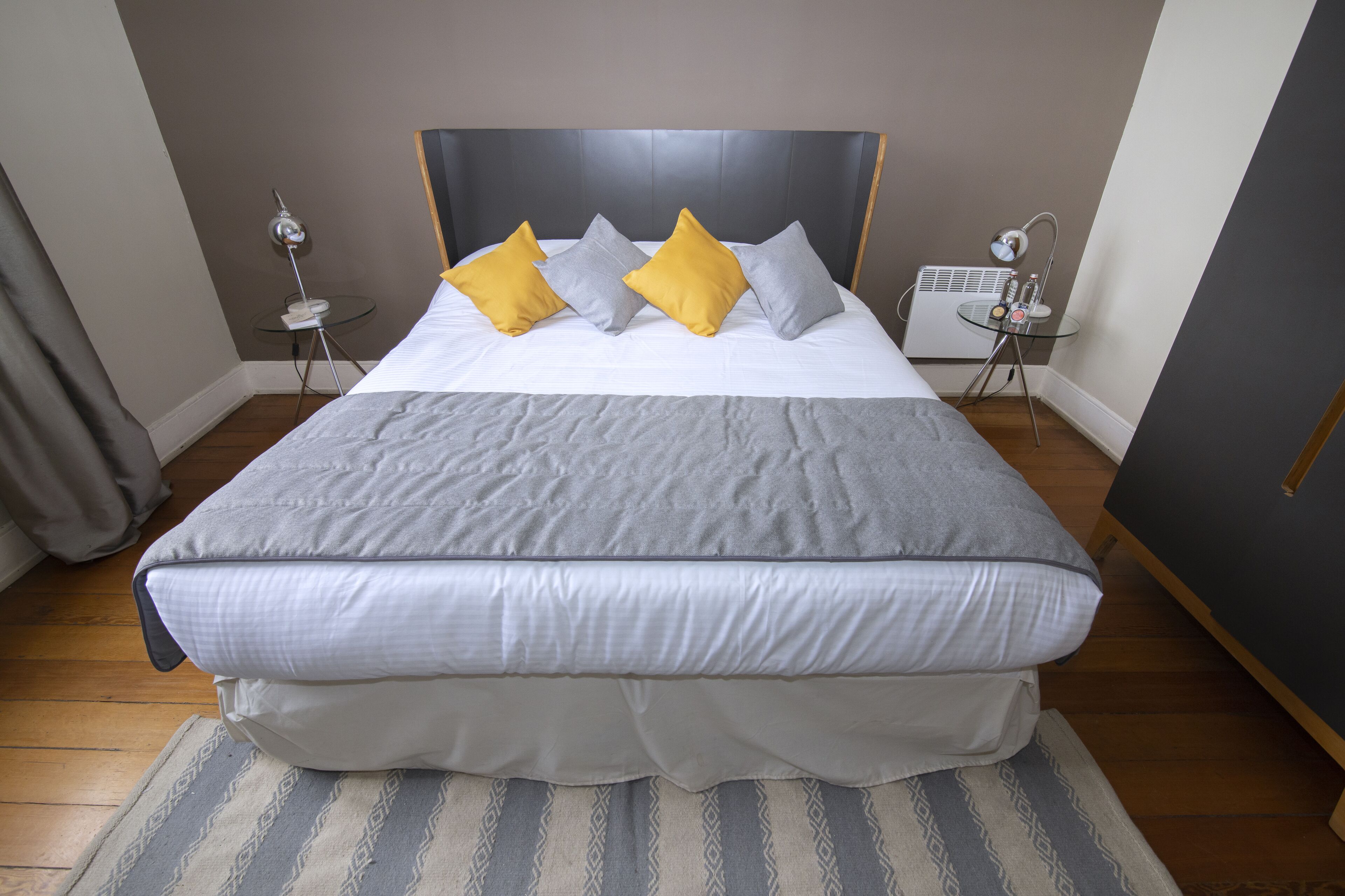 Una cama bien hecha con edredón gris y blanco, dos almohadas en amarillo y gris, una mesita de noche con una lámpara y una pequeña mesa con un radio.