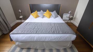 Una cama bien hecha con edredón gris y blanco, dos almohadas en amarillo y gris, una mesita de noche con una lámpara y una pequeña mesa con un radio.
