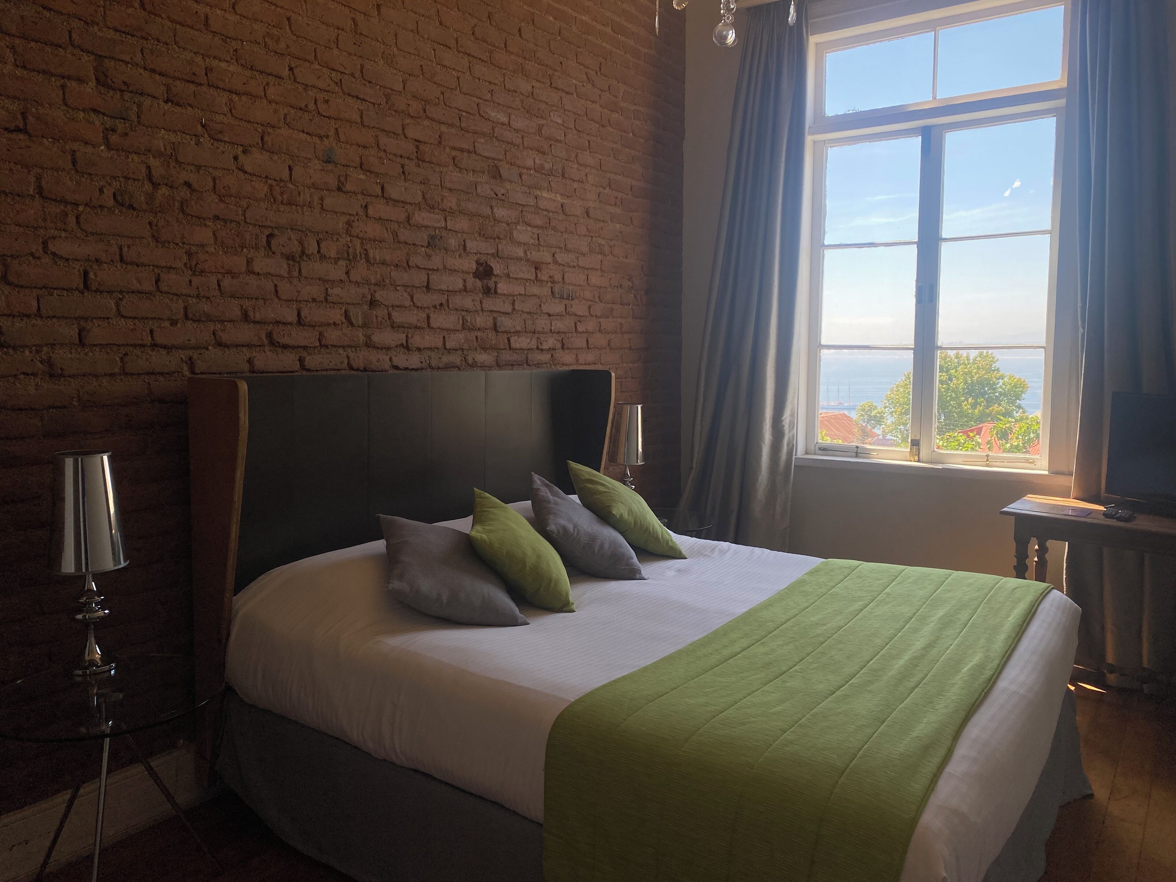 Habitación de hotel con pared de ladrillo, una cama grande con almohadas verdes y grises, una mesita de noche con lámpara y vista al mar a través de una ventana.