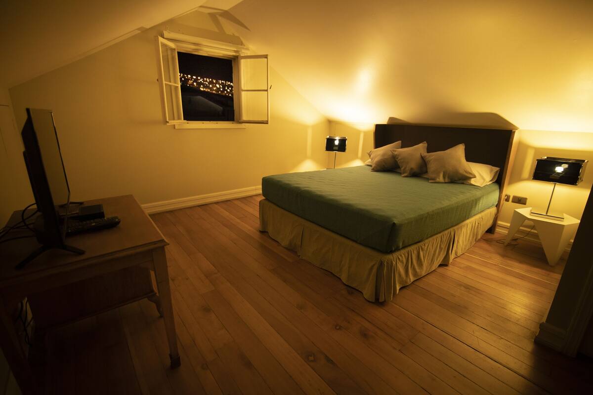 Un dormitorio con cama, escritorio, computadora, ventana y mesita de noche con lámpara.
