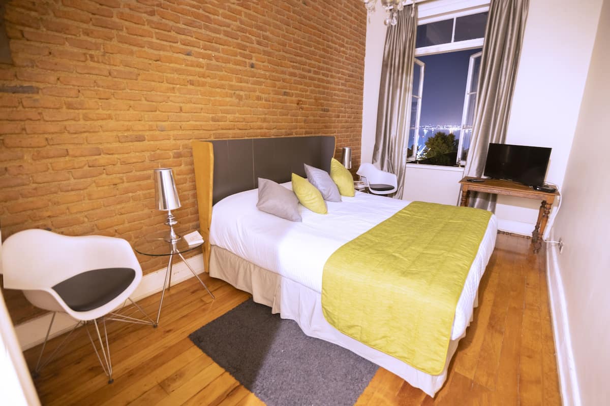 Habitación de hotel con pared de ladrillo, cama con manta verde, una silla blanca, televisor sobre un mueble de madera y ventana con cortinas.