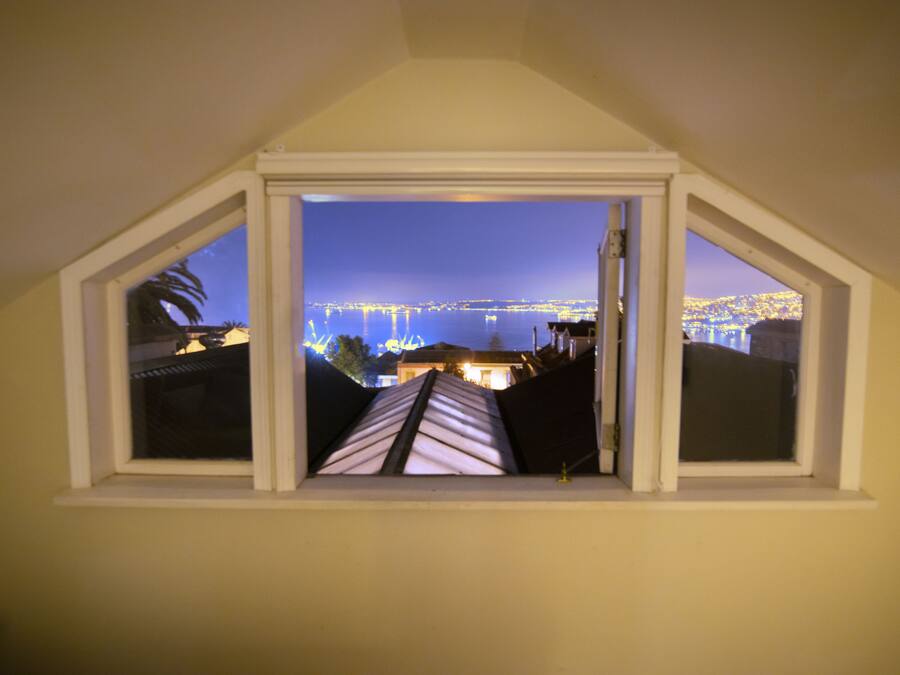 Vista da janela com moldura triangular, com vista para um horizonte urbano noturno, com edifícios iluminados e uma ponte ao longe.